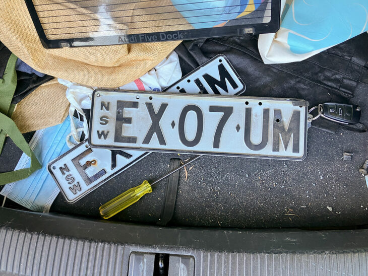 A dirty set of NSW motor vehicle number plates 'EX-07-UM' sit in the open hatch of a car.